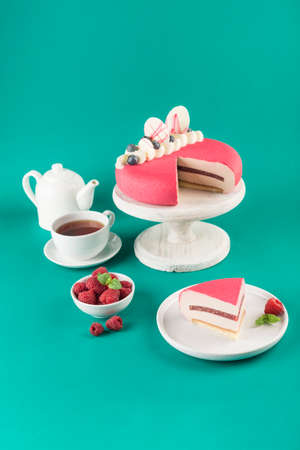 Mousse cake with a cut piece with raspberries on a wooden white stand with a teapot and a cup of tea...