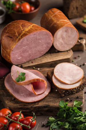 Boiled ham sliced on bread lie on a wooden board with fresh herbs and cherry tomatoes. Dark background. Side view. Vertical orientation.の写真素材
