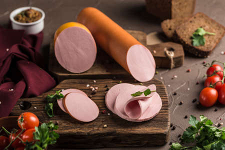 Boiled sausage cut into pieces on a wooden board with fresh herbs and cherry tomatoes on a dark background Side view. Horizontal orientation with a copy-paste for the text.の写真素材