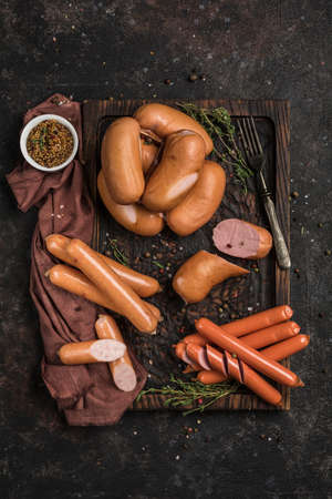 A set of different sausages on a wooden board with grainy mustard and fresh herbs on a dark background. Top view. Vertical orientation with a copy space for the text.の写真素材