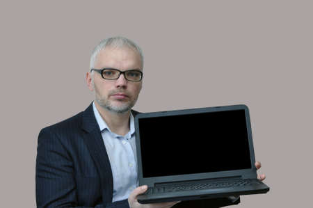 businessman in glasses and with a beard holds a laptop with a blank screen. Isolated on gray...