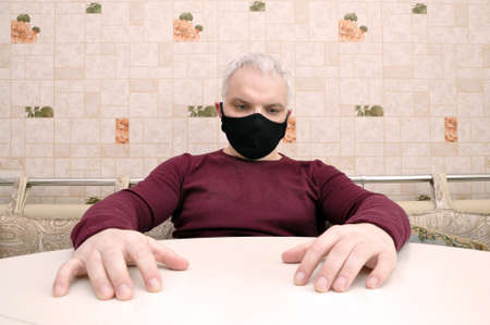 COVID-19. Stay home and distance yourself. A young man in a black medical mask is sitting at home. Try on a mask.の写真素材