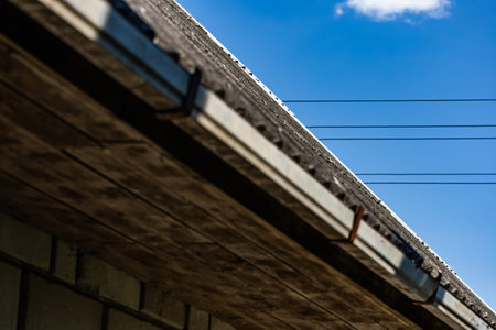 Horizontal wide angle perspective view looking up at a house roof eave, rain gutter, wooden soffit, and brick wall under a bright blue sky with power lines.