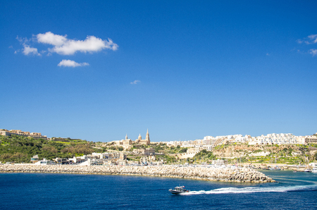 View of port village town Mgarr, catholic Ghajnsielem Parish Church and speedboat in the foreground on the Gozo island in front of blue sky with clouds, Maltaの写真素材