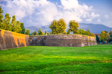 Defensive brick city wall, grass green lawn, trees and Tuscany hills and mountains with beautiful cloudy evening sky background, Lucca, Italyの写真素材