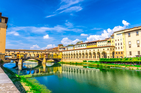Ponte Vecchio bridge with colourful buildings houses over Arno River blue reflecting water and embankment promenade archways, historical centre of Florence city, blue sky white clouds, Tuscany, Italyの写真素材