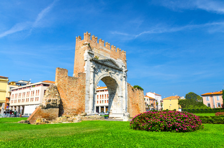 Ruins of ancient brick wall and stone gate Arch of Augustus Arco di Augusto, green lawn with bush of flowers in old historical touristic city centre Rimini, blue sky background, Emilia-Romagna, Italyの写真素材