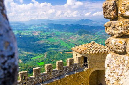 Aerial top panoramic view landscape with valley, green hills, fields, villages of Republic San Marino suburban district, blue sky white clouds background and stone fortress wall and tower with merlonsの写真素材