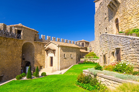 Courtyard with green grass lawn of Prima Torre Guaita first medieval tower with stone brick fortress wall with merlons on Mount Titano rock, blue sky white clouds background, Republic San Marinoのeditorial素材