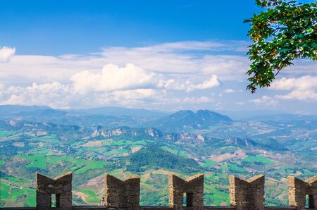 Aerial top panoramic view landscape with valley, green hills, fields and villages of Republic San Marino suburban district with blue sky white clouds background and stone fortress wall with merlonsの写真素材