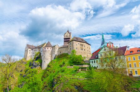 Loket Castle Hrad Loket gothic style building on massive rock and colorful buildings in Loket town, green trees and hills, blue cloudy sky background, Karlovy Vary Region, West Bohemia, Czech Republicのeditorial素材