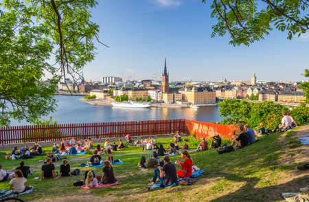 Sweden, Stockholm, May 29, 2018: Young people spend time (sit, drink, eat) on lawn with green grass and trees around on Sodermalm island with view of old historical town Riddarholmen and Gamla Stanのeditorial素材