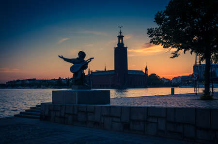 Sweden, Stockholm, May 29, 2018: Silhouette of Evert Taube statue monument and City Hall Stadshuset Municipal Council on Kungsholmen Island at sunset, dusk, twilight, blue yellow orange sky backgroundのeditorial素材
