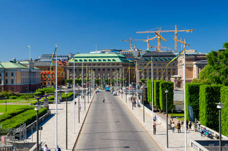 Sweden, Stockholm, May 31, 2018: Riksplan green grass lawn, bushes and street with national flags on Sodermalm island, Gustav Adolfs torg, Royal Swedish Opera house, blue sky with yellow cranesのeditorial素材