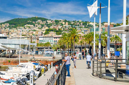 Genoa, Italy, September 11, 2018: man is riding bicycle on promenade embankment and people are looking at white yachts in marina harbour of Genova with beautiful view of colorful villas on green hillのeditorial素材