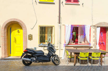 Rimini, Italy, September 19, 2018: table with chairs and motorcycle bike scooter parked on cobblestone street near buildings with colorful wall, door and windows in historical city centreのeditorial素材