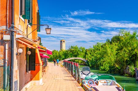 Torcello island with embankment along water canal with motor boats and buildings along promenade. Stone Devil bridge, green trees and tower background. Venetian Lagoon, Veneto Region, Italy.の写真素材