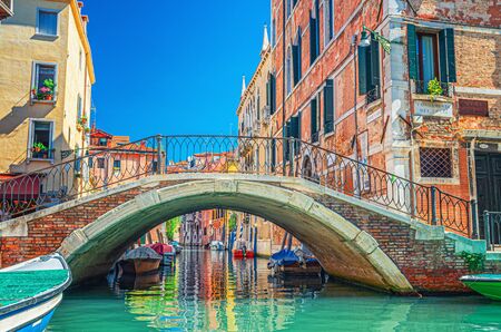 Bridge across narrow water canal in Venice with moored boats between old colorful buildings with balconies and brick walls, blue sky, Veneto Region, Northern Italy. Typical Venetian cityscapeの写真素材