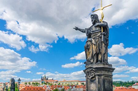 Saint John the Baptist statue on Charles Bridge Karluv Most over Vltava river with Prague Castle, St. Vitus Cathedral in Hradcany district, blue sky white clouds background, Bohemia, Czech Republicの写真素材