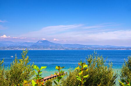 Garda lake with blue azure turquoise water and wooden pier dock, coast with mountain range, blue sky background, Sirmione town, Lombardy, Northern Italyの写真素材