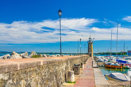 Stone pier mole with lighthouse, street lights and yachts on boat parking port marina in Desenzano del Garda town, blue sky white clouds background, Lombardy, Northern Italyの写真素材
