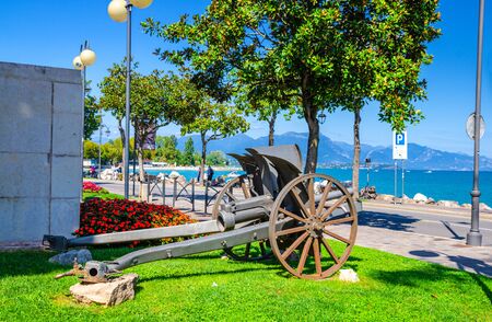 Cannon on green grass lawn in historical centre of Desenzano del Garda town with view of Garda Lake promenade embankment and mountain range, blue sky background, Lombardy, Northern Italyの写真素材