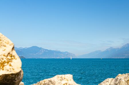 Small sailing yacht on Garda Lake azure water with Monte Baldo mountain range, blue sky white clouds background, Desenzano del Garda town, view from stone pier mole, Lombardy, Northern Italyの写真素材