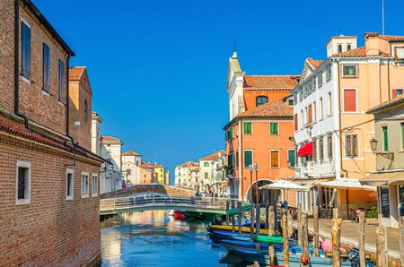 Chioggia cityscape with narrow water canal Vena with moored multicolored boats between old colorful buildings and brick bridge, blue sky background in summer day, Veneto Region, Northern Italyの写真素材