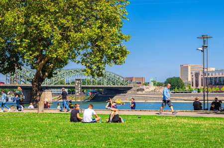 Cologne, Germany, August 23, 2019: people tourists walking down and having a rest on green grass lawn near Rhine river promenade in historical city centre, Hohenzollern Bridge and blue sky backgroundのeditorial素材