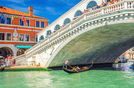 Venice, Italy, September 13, 2019: gondolier on gondola traditional boat sailing on water of Grand Canal waterway under Rialto Bridge, typical colorful buildings and clear blue sky backgroundのeditorial素材