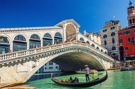 Venice, Italy, September 13, 2019: gondolier on gondola traditional boat sailing on water of Grand Canal waterway under Rialto Bridge, typical colorful buildings and clear blue sky backgroundのeditorial素材