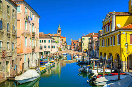 Chioggia cityscape with narrow water canal Vena with moored multicolored boats between old colorful buildings and brick bridge, blue sky background in summer day, Veneto Region, Northern Italyの写真素材