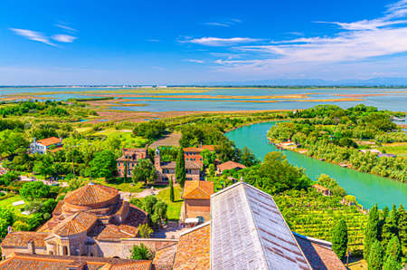 Aerial view of Torcello islands with buildings, water canal, swamp, green trees and bushes. Panoramic view of Venetian Lagoon from bell tower. Veneto Region, Northern Italy. Blue sky background.の写真素材