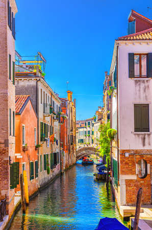Venice cityscape with narrow water canal with boats moored between old colorful buildings and stone bridge, Veneto Region, Northern Italy. Typical Venetian view, vertical view, blue sky backgroundの写真素材