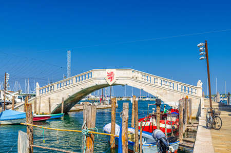 Stone bridge Ponte di Vigo across Vena water canal with colorful boats near pier embankment in historical center of Chioggia town, blue sky background in summer day, Veneto Region, Northern Italyの写真素材