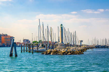 Lighthouse on stone pier of yacht port marina, wooden bricole poles in water of lagoon near Chioggia town, blue sky in summer day, Veneto Region, Northern Italyの写真素材