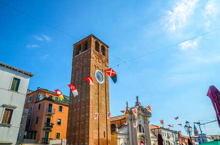 Chioggia, Italy, September 16, 2019: Chiesa Saint Andrea catholic church building with brick bell tower and medieval small flags in historical town center, blue sky in summer day, Veneto Regionのeditorial素材