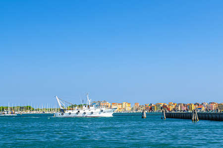 Chioggia, Italy, September 16, 2019: fishing ship boat in water of lagoon with row of colorful buildings of Sottomarina town, blue sky background in summer day, Veneto Regionのeditorial素材