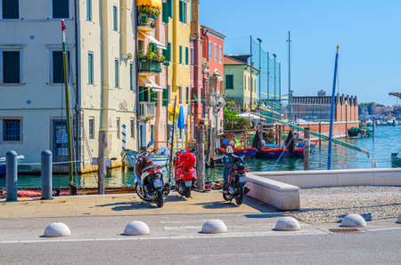 Chioggia, Italy, September 16, 2019: motorcycles and bikes vespa on embankment, old buildings and boats in water in port marina in historical town center, blue sky in summer day, Veneto Regionのeditorial素材