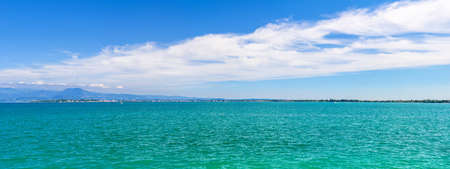 Panorama of Garda Lake azure turquoise water with view of Monte Baldo mountain range and Sirmione peninsula, blue sky white clouds background, Desenzano del Garda town, Lombardy, Northern Italyの写真素材