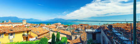 Panorama of Desenzano del Garda town with red tiled roof buildings, Garda Lake water, Monte Baldo mountain range, Sirmione peninsula, Lombardy, Northern Italy. Aerial panoramic view of Desenzanoの写真素材