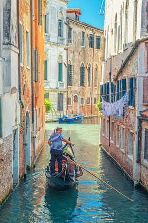 Venice, Italy, September 13, 2019: Gondola sailing narrow canal between old buildings with brick walls. Gondolier dressed traditional white and blue striped short-sleeved polo shirt and boater hatのeditorial素材