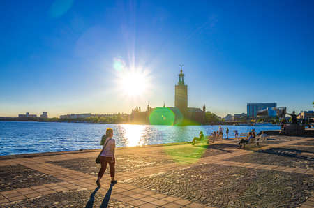 Sweden, Stockholm, May 30, 2018: People are sitting on bench at promenade embankment of Lake Malaren looking at Stockholm City Hall Stadshuset tower building on Kungsholmen Island, view against sunのeditorial素材