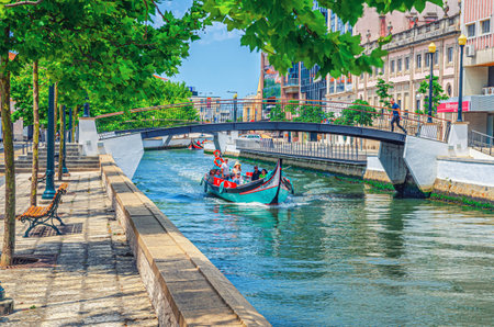 Aveiro, Portugal, June 13, 2017: traditional colorful Moliceiro boat with tourists sailing in narrow water canal, bridge across waterway and buildings along embankment promenade in city centerのeditorial素材