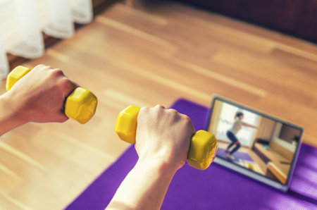 Young woman hands raising yellow dumbbells during workout sports at home, video exercises tutorials on tablet screen on floor mat in living room background, close-up view from back, sport conceptの写真素材