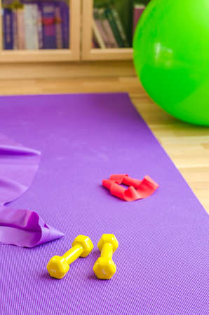 Sports equipment on floor in living room, violet yoga mat, yellow dumbbells, red rubber resistance band and green rubber fitness aerobic ball, bookcase background, home workout concept, vertical viewの写真素材