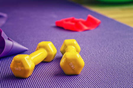 Sports equipment close-up view, yellow dumbbells and red rubber resistance band on violet ribbed yoga mat, home workout conceptの写真素材