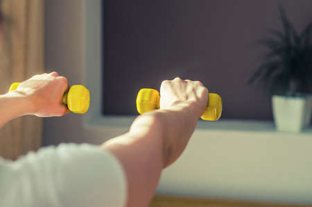 Young woman arms raising yellow dumbbells during workout sports at home, flower pot with green plant in wall niche in living room background, close-up view from back, sun light from sideの写真素材