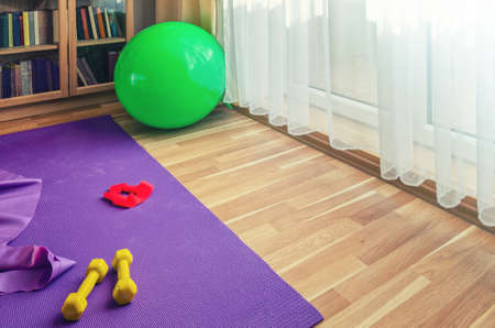 Sports equipment on floor in living room, violet yoga mat, yellow dumbbells, red rubber resistance band and green rubber fitness aerobic ball, bookcase near window background, home workout conceptの写真素材