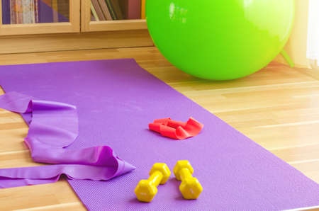 Sports equipment on floor in living room, violet yoga mat, yellow dumbbells, red rubber resistance band and green rubber fitness aerobic ball, bookcase background, home workout conceptの写真素材